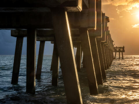Morning sunlight cuts through the underside of Juno Beach Pier, creating dramatic beams of light and deep shadows between the wooden pilings.