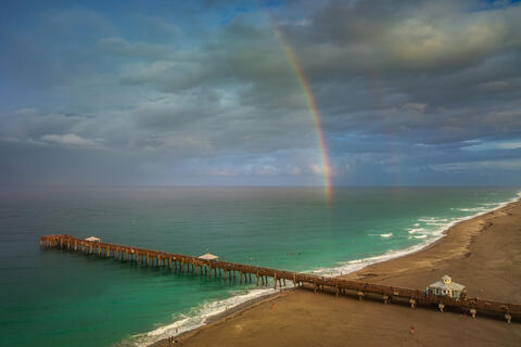 Aerial view of a rainbow arching over the turquoise shoreline near Juno Beach Pier, with the beach, pier, and sea visible from above.