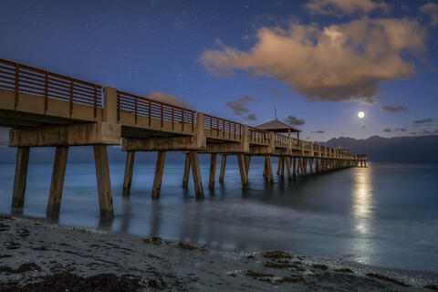 Juno Beach Pier stands beneath a cool blue night sky with the moon resting softly above the horizon over a calm, still ocean.