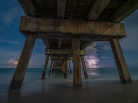 A bolt of lightning tears across the dark sky beyond Juno Beach Pier, illuminating the ocean and storm clouds in a dramatic instant.