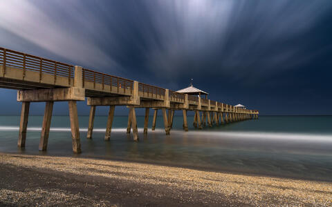 Long exposure photograph of heavy storm clouds and streaking skies gathering over Juno Beach Pier, with softened waves below.