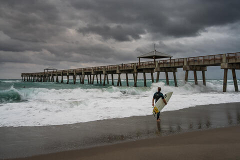A lone surfer walks along the shoreline beside Juno Beach Pier as dark storm clouds build overhead and waves roll in with force.