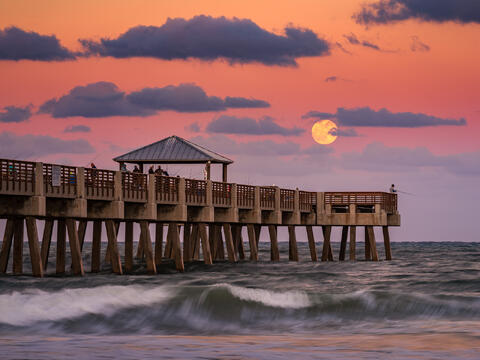 The moon rises above the horizon beside Juno Beach Pier as warm evening clouds catch the last traces of color at dusk.