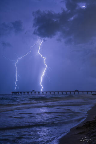 Lightning bolts illuminate a dark ocean horizon with a silhouette of a pier in the foreground during a nighttime thunderstorm.