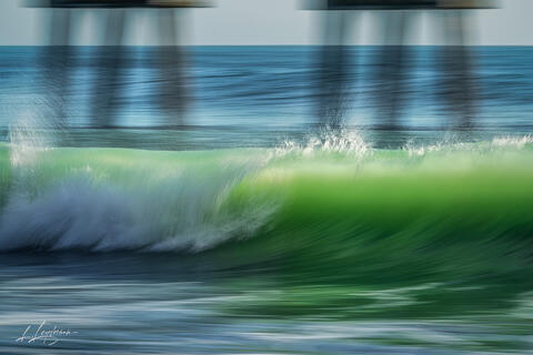 A wave under a pier rendered in motion, creating emerald-green strokes like a painting on nature’s canvas.