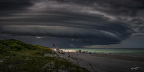 A dramatic storm cloud presses low over the Florida coast as a narrow beam of light breaks through the darkness. The ocean and beach below sit in heavy silence,