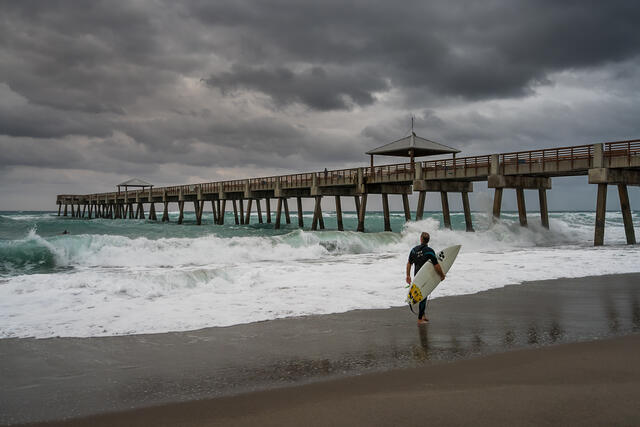 A lone surfer walks along the shoreline beside Juno Beach Pier as dark storm clouds build overhead and waves roll in with force.