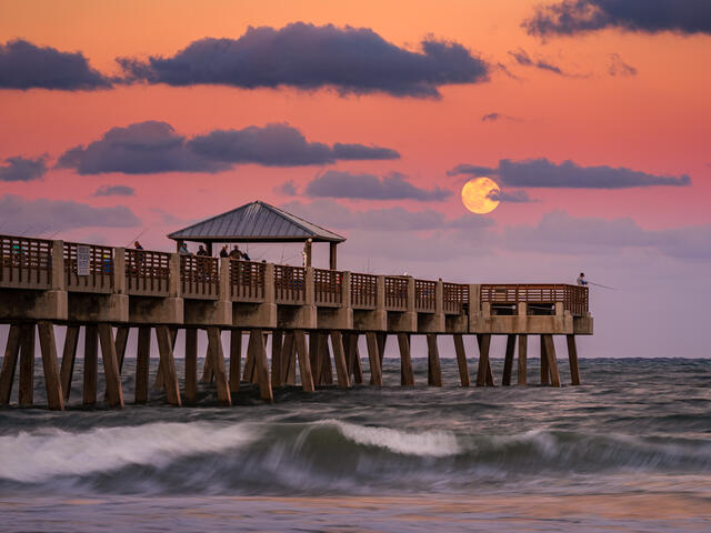 The moon rises above the horizon beside Juno Beach Pier as warm evening clouds catch the last traces of color at dusk.