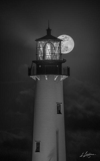A black-and-white photo of a lighthouse with a glowing full moon perfectly aligned behind its lantern, captured at night in a calm and dramatic seascape scene.