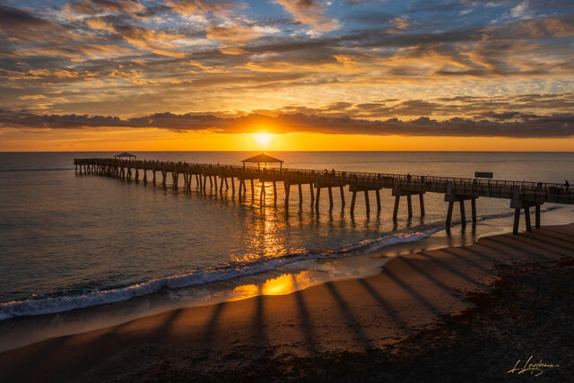 Aerial view of Juno Beach Pier at sunrise with dramatic golden light casting long diagonal shadows across the wet sand, surrounded by ocean waves and vibrant or