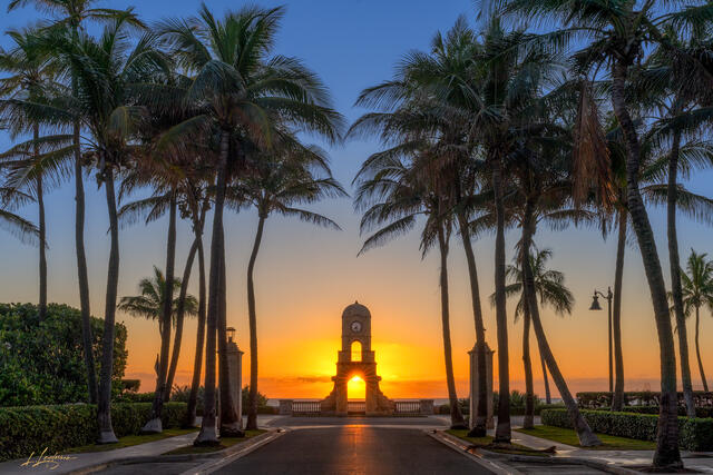 Sunrise framed inside the Worth Avenue clock tower in Palm Beach, Florida, with tall palm trees lining the road and golden light over the ocean.